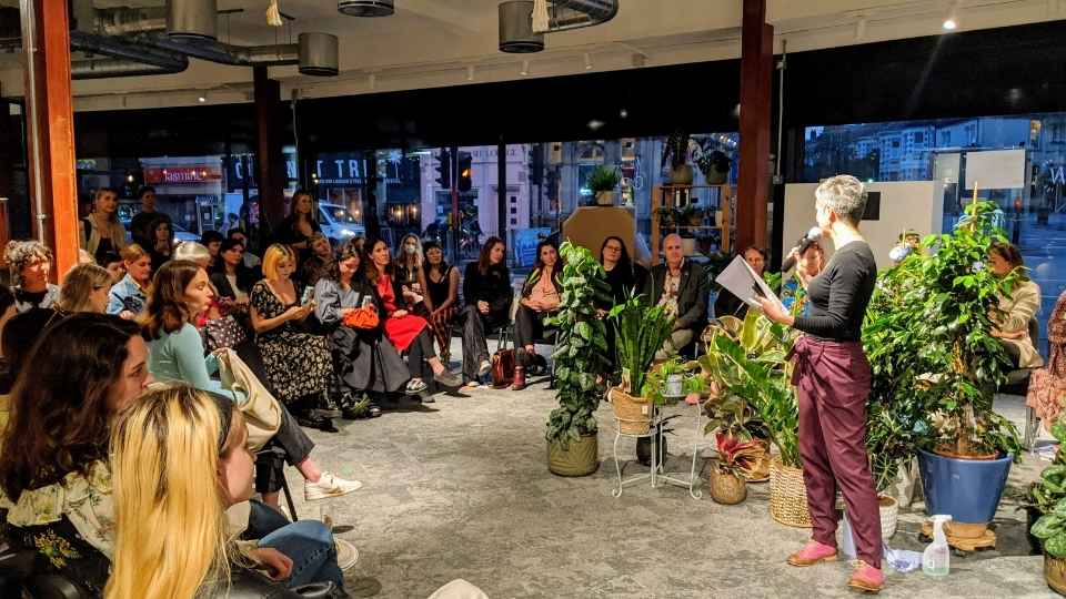 Katherine Piper speaking to the Fashion Show audience in the round surrounded by brightly dressed audience members and plants at The Future Leap Co-working Hub