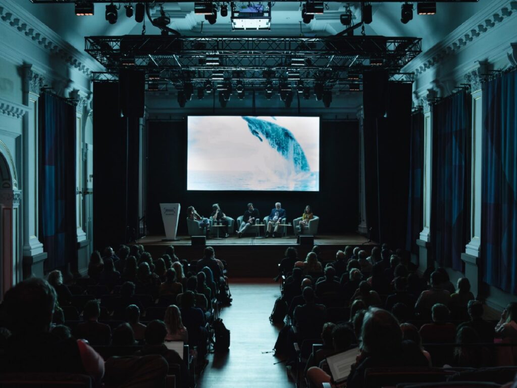 A seated audience watching a film being projected above a panel of speakers on a stage. The film depicts a whale jumping out of the ocean.