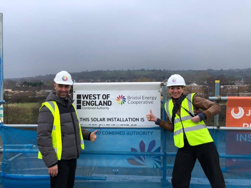 two people with yellow high vis and hard hats stand on a roof of solar panels for Bristol Energy Cooperative